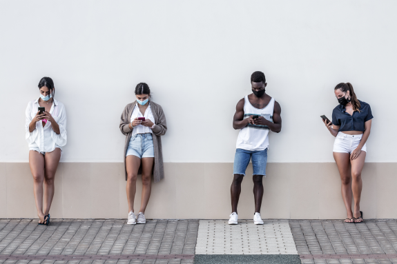 Four people wearing masks standing apart using smartphones against a wall.