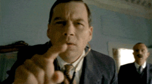 Man in a suit pointing at the camera indoors with another person in the background.