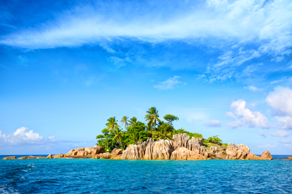 Small tropical island with palm trees and rocks in a bright blue ocean under a clear sky.