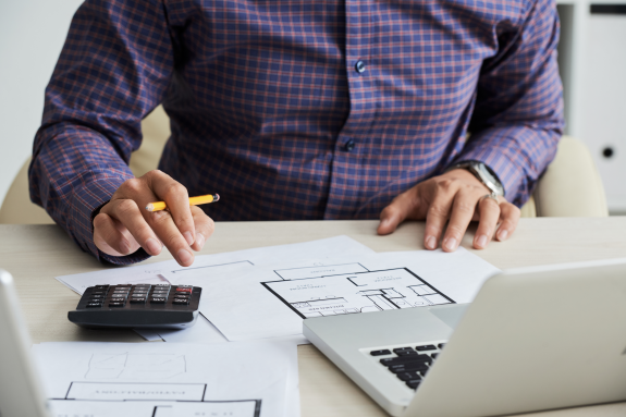 Person in plaid shirt working on blueprints with calculator and laptop on desk