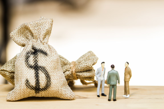 Miniature businessmen figures near burlap money bags with dollar signs.
