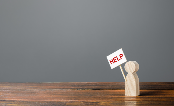 Wooden figure holding a help sign on a wooden table with gray background.
