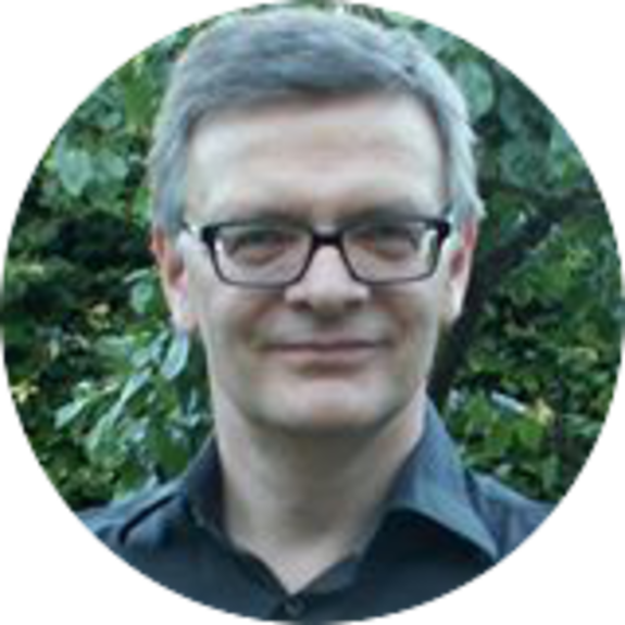 Man wearing glasses and a dark shirt smiling outdoors with greenery in background.