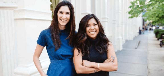 Two women smiling and standing outdoors with an urban background.